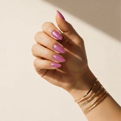 Hand wearing Delune Glitter Bug pink glitter nail wraps with gold bangles against a warm plaster wall